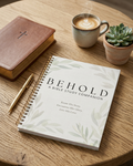 A spiral-bound Bible study companion book titled 'BEHOLD' lies on a round wooden table, surrounded by a leather-bound Bible, a cup of coffee with latte art, and a small potted succulent.