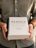 A person in a muted green button-up shirt holds a stack of spiral-bound books, with the top book titled “BEHOLD: A BIBLE STUDY COMPANION,” against a softly blurred neutral background.