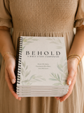 A person in a beige dress holds a stack of spiral-bound Bible study companion books titled "BEHOLD: A BIBLE STUDY COMPANION" with a white cover and soft green botanical illustrations.