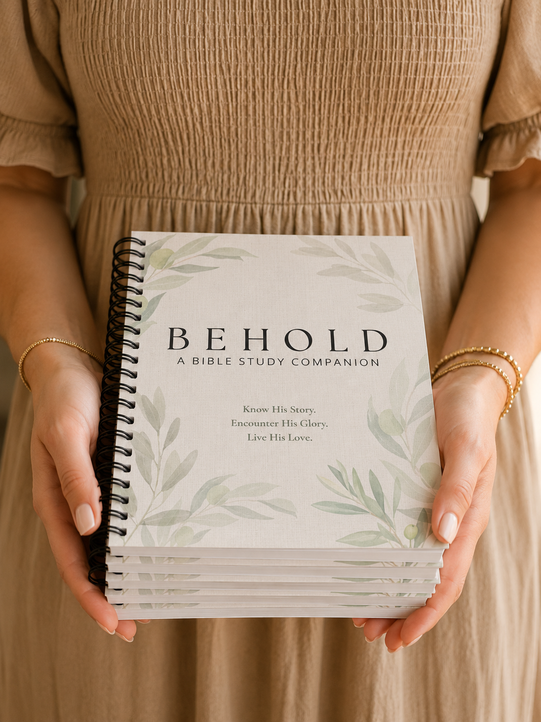 A person in a beige dress holds a stack of spiral-bound Bible study companion books titled "BEHOLD: A BIBLE STUDY COMPANION" with a white cover and soft green botanical illustrations.